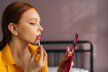 Closeup face of sexy young woman with red hair applying lipstick holding mirror and looking at herself. Close up of beauty female looking facial at mirror for makeup cosmetic with lips elegance.