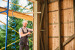 Carpenter constructing wooden framed house. Bearded man worker cladding facade of house, fastening with screwdriver, wearing work overalls and helmet. Concept of modern eco-friendly construction.