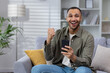 © Tetiana - Portrait of a happy young African American man sitting on the couch at home and looking at the camera while using the phone. Celebrating victory, winning