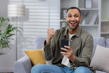 Portrait of a happy young African American man sitting on the couch at home and looking at the camera while using the phone. Celebrating victory, winning