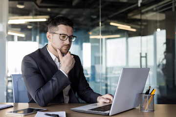 Wall Mural - Concentrated and serious young man businessman works in the office at a laptop on a new project, looks intently at the monitor, solves a question, conducts an online conference