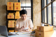 Young asian sme businessman working at warehouse, home office, independent courier company with parcel boxes, checking customer address with laptop.