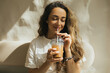 © polinaloves - Young woman with long curly hair drinking iced coffee on beige background.