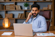 © Ivan - Handsome indian business man entrepreneur sitting at work desk talking on cell phone making telephone mobile call by cellphone working at home or in office.