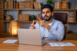 © Ivan - Millennial arab freelancer man in casual sitting at work desk in front of laptop, gesturing and smiling, having video call with colleagues while working from home, copy space