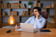 © Ivan - Handsome arab man with beard sits at work desk with laptop, talk by phone, looks at empty space in living room interior. Work at home, social networks, call to client remote, great offer and ad