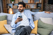 © Ivan - young man holding using cell phone sitting on sofa couch in living room. Cool guy browsing internet, surfing web