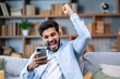 © Ivan - Excited overjoyed young man sit on sofa holding smartphone feeling euphoric with mobile online bet gambling victory. Ecstatic guy looks at cellphone scream with joy celebrate fantastic news received