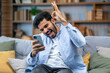 © Ivan - Surprised happy man holding phone looking at cellphone read good news in sms sit on sofa, amazed guy winner excited by mobile app win scream with joy celebrate victory success at home
