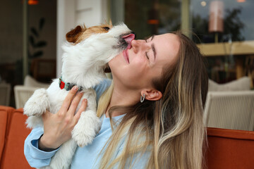 Wall Mural - Wire haired jack russell terrier licking a cheek of his joyful woman outside of the coffee shop. Young woman and her dog playing outdoors. Copy space, background.