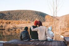 Family Wearing Casual Knit Clothes Resting At Wooden Deck Over Lake And Autumn Nature Background. Fall Season. Parenthood. Back View.