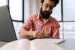 © dikushin - Closeup low-angle view portrait of bearded Indian businessman using laptop analyzing working project results, writing notes siting at table by window. Focused student male learning remotely.