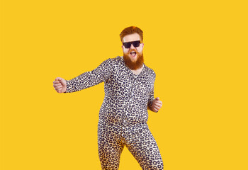 Funny eccentric overweight man in pajamas with leopard print having fun, dancing and fooling around. Millennial red-bearded fat man in pajamas and black glasses dances isolated on orange background.