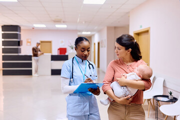 Wall Mural - Black nurse writes data in medical record while talking to mother with baby at doctor's office.