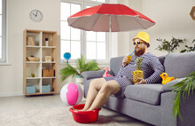 Funny Man In Bathing Suit, Sunglasses And Bright Yellow Panama Hat Is Sitting At Home On The Sofa In The Living Room Under Umbrella, Imagining, Dreaming About Vacation At The Sea, Drinking Cocktail.