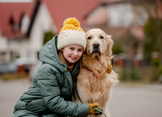 Wall Mural - Preteen child girl hugging golden retriever dog at autumn city street wearing hat and warm jacket. Pretty kid with purebred pet doggy labrador outdoors