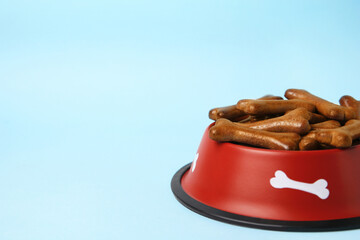 Wall Mural - Red bowl with bone shaped dog cookies on light blue background, closeup. Space for text