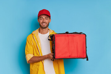 Friendly delivery guy in red cap and yellow jacket holding red delivery box and smiling, posing against blue backdrop, fast delivery concept, copy space