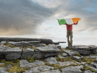 Man with National flag of Ireland in rough stone field of a cliff, Aran island, county Galway, Ireland. Travel and tourism concept. Stunning nature scenery in the background.