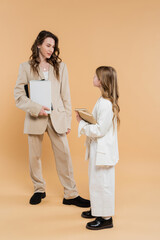 Wall Mural - modern family, mother and daughter in suits, woman and child holding books and notebooks while standing together on beige background, formal attire, corporate mom, education concept