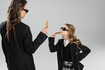 Wall Mural - mother and daughter in sunglasses, businesswoman in suit and schoolgirl in uniform holding credit cards and looking at each other on grey background, modern parenting, money management