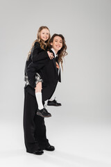 Wall Mural - cheerful mother piggybacking her daughter, working mom in business attire and schoolgirl in uniform on grey background in studio, modern parenting, fashionable family, having fun