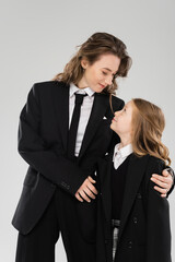 Wall Mural - modern parenting, cheerful businesswoman in suit hugging daughter in school uniform and standing together on grey background, happy mother and child, back to school