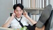 © M Stocker - Young asian man putting feet on table in office while enjoy talking by smartphone take break from work. Happy businessman cellphone