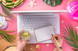 Summer office, working background, blogger workplace flat lay. White laptop keyboard with notebook, straw hat, tropical decor and leaves, cold cocktail glass on high-colored sunny pink background