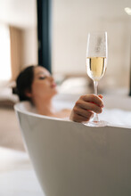 Vertical Selective Focus Shot Of Young Relaxed Woman Lying In Foam Bath Holding Glass Champagne Enjoying Alcoholic Drink During Luxurious Recreation For Weekend. Brunette Female Relaxing In Bathroom.