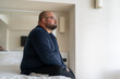 © DimaBerlin - Sad fat lonely man sitting on bed in hotel and looking out window, thinking about life problems, no strength change, side view overweight depressed man no friends and mental disorders