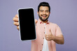© ViDi Studio - Young Indian man wears pink shirt white t-shirt casual clothes hold in hand use close up mobile cell phone with blank screen workspace show thumb up isolated on plain pastel light purple background.