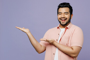 Young Indian man he wear pink shirt white t-shirt casual clothes point hands arms aside indicate on workspace area copy space mock up isolated on plain pastel light purple background studio portrait.