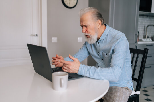 Side view of angry bearded mature elderly man with grey hair listening to companion during video call on laptop and can barely contain himself from swearing, sitting at desk with coffee in kitchen