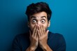 © Markus Schröder - Medium shot portrait photography of a glad boy in his 30s making a surprise gesture by covering one's mouth against a sapphire blue background. With generative AI technology