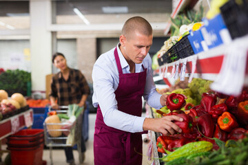 Sticker - Supermarket worker stacking red peppers on shelf in salesroom. Woman with cart shopping in background.