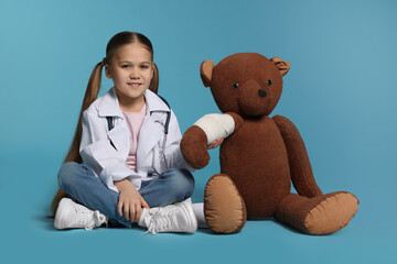 Wall Mural - Little girl in medical uniform and toy bear with bandage on light blue background