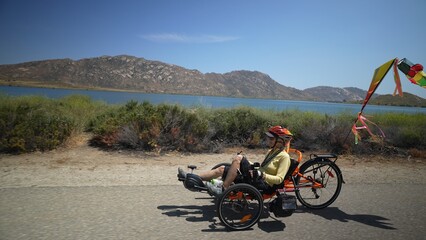 Slow motion of elderly senior woman riding e-bike recumbent tricycle bike on a path on a sunny day beside a lake.