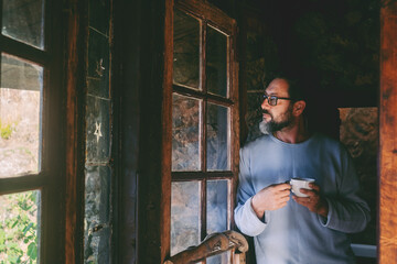 one man standing at home chalet drinking tea and looking outside the window with serious thoughtful 