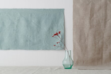 Horizontal shot of minimalistic still life installation with glass vessel and dried red flowers against white wall, gray and blue linen fabric background, copy space
