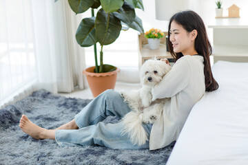 Asian young happy cheerful female owner sitting smiling on fluffy carpet floor holding hugging cuddling embracing best friend companion dog white long hair mutt shih tzu together in bedroom at home