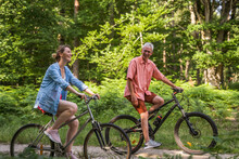 Happy Senior Father Riding Bicycles With His Daughter Through The Path In Green Forest