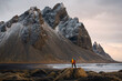 A tourist couple stands on the beach, admiring the majestic reflection of the Vestrahorn mountain at Cape Stokksnes, Iceland, as the sun rises on the horizon. The beautiful snow-capped mountains.