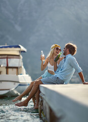 Wall Mural - Couple having a beer and laughing together sitting by water. Summertime holiday. Togetherness, lifestyle, luxury concept.