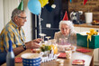 © luckybusiness - Happy senior woman blowing candles on her birthday cake, sitting at table with husband cheering her.