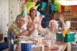 © luckybusiness - Happy family of senior couple and daughter cheering for birthday senior woman, celebrating her birthday together, together in the home kitchen by dinner table.