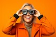 © Markus Schröder - Close-up portrait photography of a happy mature woman imitating the use of binoculars with the hands against a bright orange background. With generative AI technology