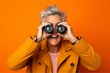 Close-up portrait photography of a happy mature woman imitating the use of binoculars with the hands against a bright orange background. With generative AI technology