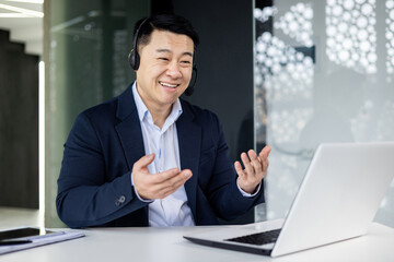 A young handsome Asian businessman in headphones with a microphone sits at a laptop in the office, conducts an online conference, a remote meeting with clients, partners. Tells, explains, smiles.