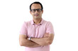 © Stella - Portrait of a middle aged happy smiling Indian man in a pink T-shirt wearing glasses, standing with arms crossed isolated on white background, confident Asian male smiling and keeping arms crossed.